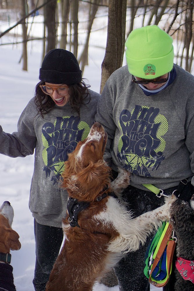 Two women wearing "I'm with the Pack" Sweatshirts walking 3 dogs in a snowy forest