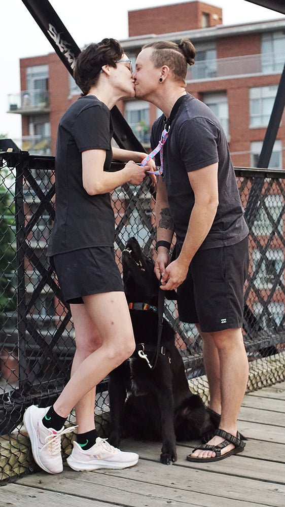 Two people kissing while one of them wears a 2023 Pride Leader attached to a seated black dog on a bridge