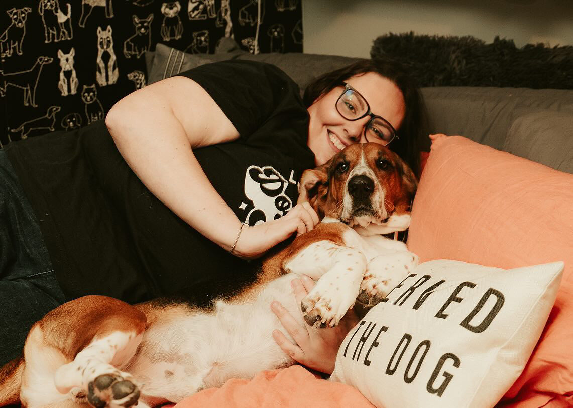 A causcasian woman in a black shirt reclines with her bassethound on a couch. There are coral pink pillows and sheets.