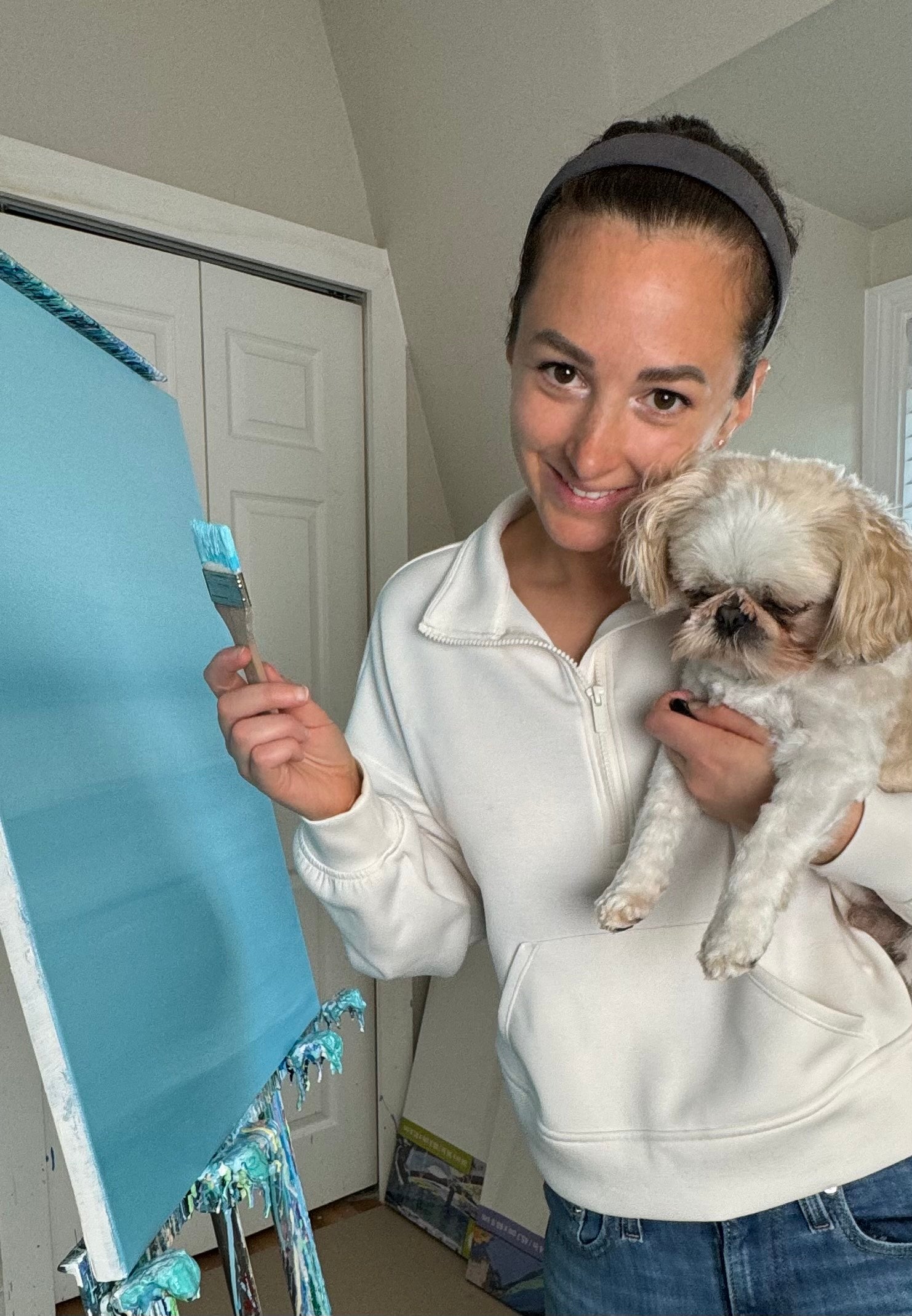 A Caucasian woman holds a small white and tan dog in her left arm. With the right arm she is painting a canvas with a light blue, abstract design.